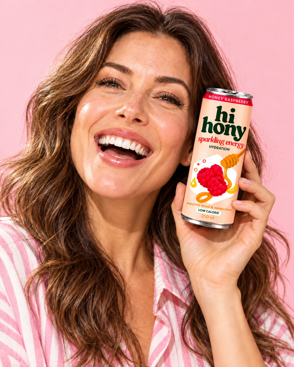 A smiling woman holding the peach HI HONY Honey Raspberry can in a playful pink campaign portrait